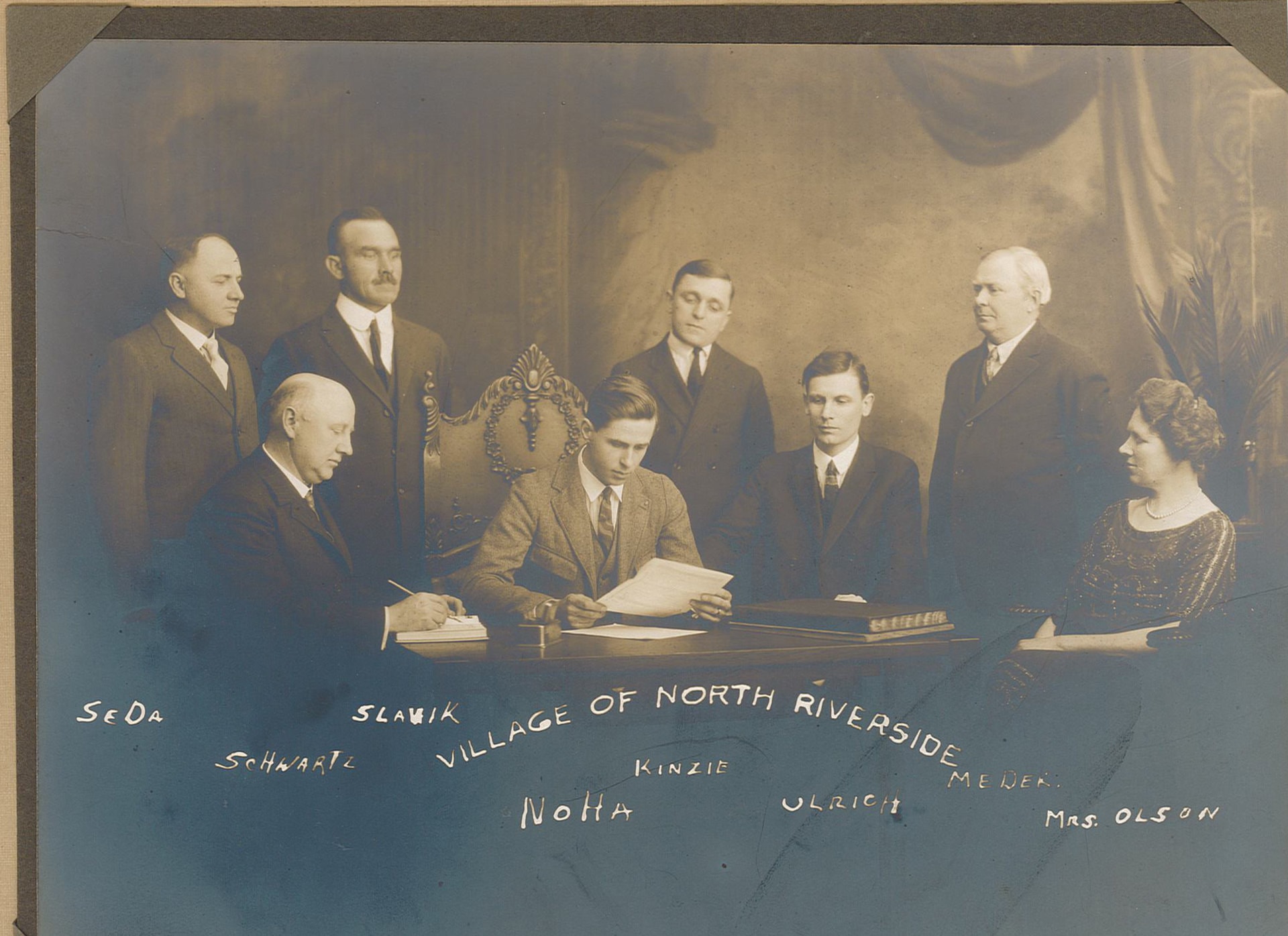 Village of North Riverside Founders picture with the names: SeDa, Schwartz, Slavik, Noha, Kinzie, Ulrich, Meder, Mrs. Olson. There are 7 men and 1 women seated and standing around a table.