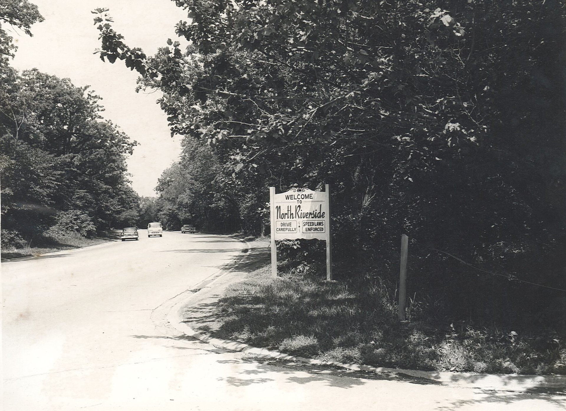 Old image of North Riverside welcome sign with a windy paved road and 3 cars. The sign reads: Welcome to North Riverside. Under that in 2 boxes are: Drive Carefully; Speed Laws Enforced.